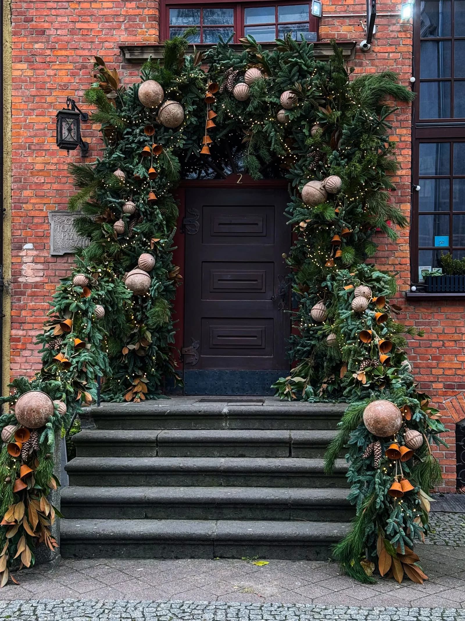 Grand evergreen garland arch with bells and ornaments framing a dark wood door on a brick building