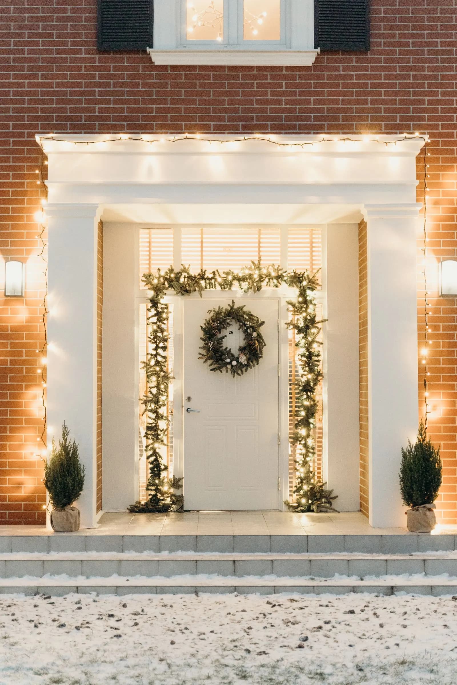 White portico entry with garland framing the door, an evergreen wreath, and warm string lights at dusk